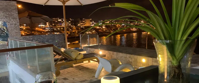 Terrasse éclairée le soir, avec chaise longue et parasol, face aux lumières des immeubles qui bordent la baie